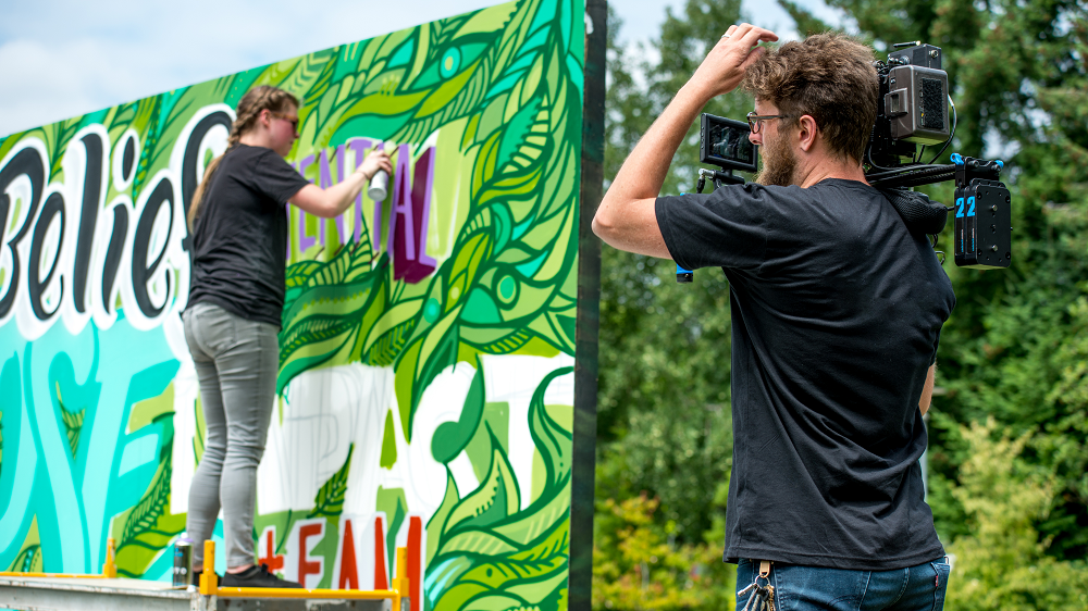 Male camera operator filming a woman standing on a ladder spray painting a wall outside