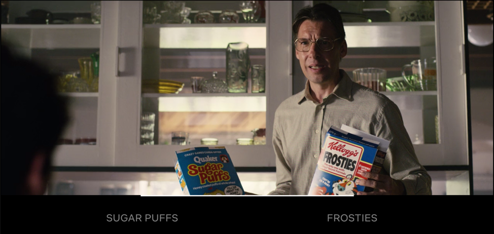 Middle-aged man with glasses holding a box of Quaker Sugar Puffs cereal in one hand and Kellogg's Frosties in the other hand.