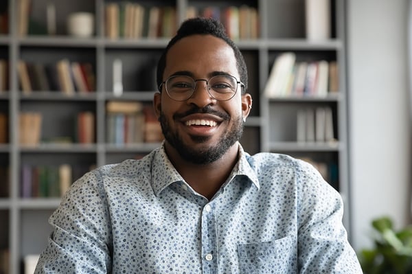 Man wearing glasses smiling at a webcam with a bookcase in the background 