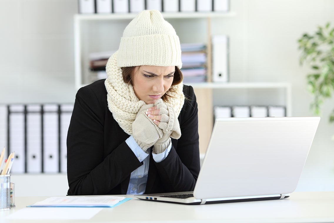 A woman looking cold wearing a beanie, scarf, and gloves while on her computer in a clean office workspace