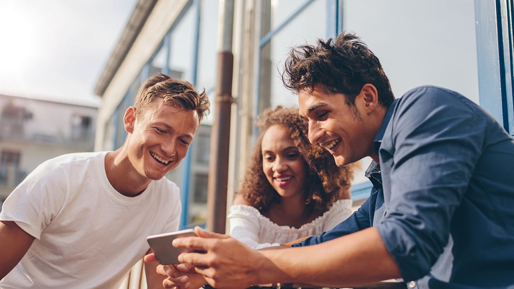 Three young friends sitting outdoors and looking at mobile phone. Group of people sitting at outdoor cafe and watching video on the smartphone.