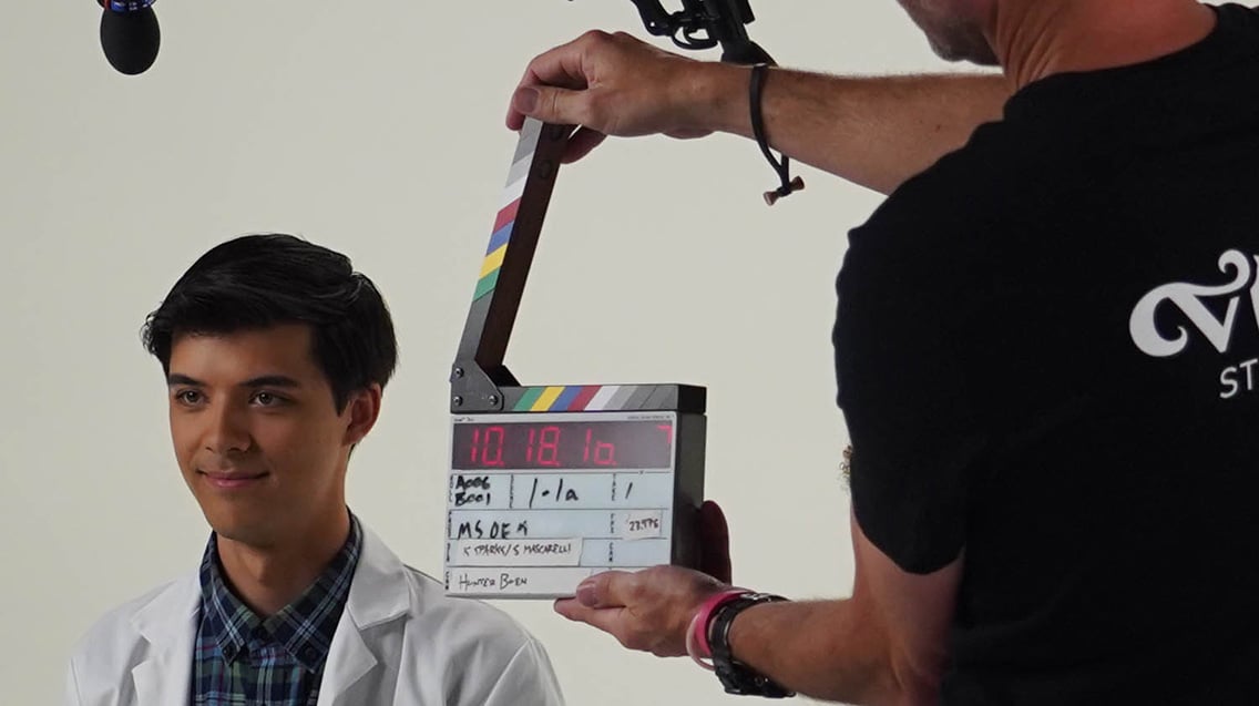 Person holds a digital clapper board with synced timecode near on-camera talent during a video production shoot in a studio