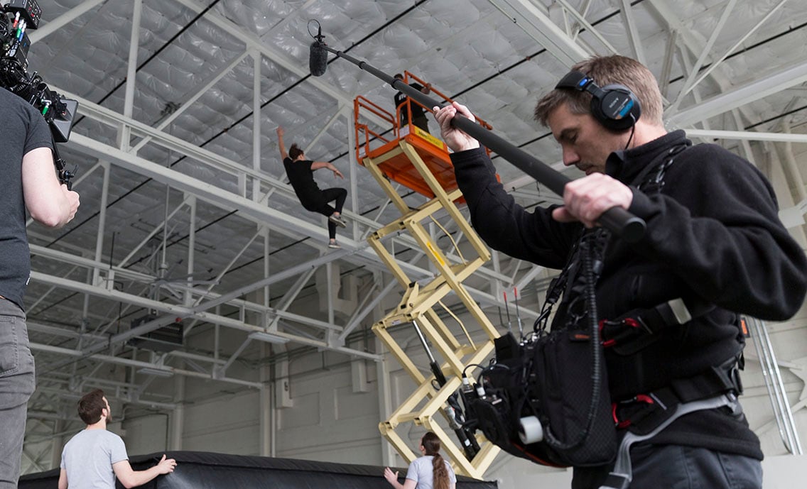 Sound tech operating the boom during a video production shoot with a woman falling from a crane in the background